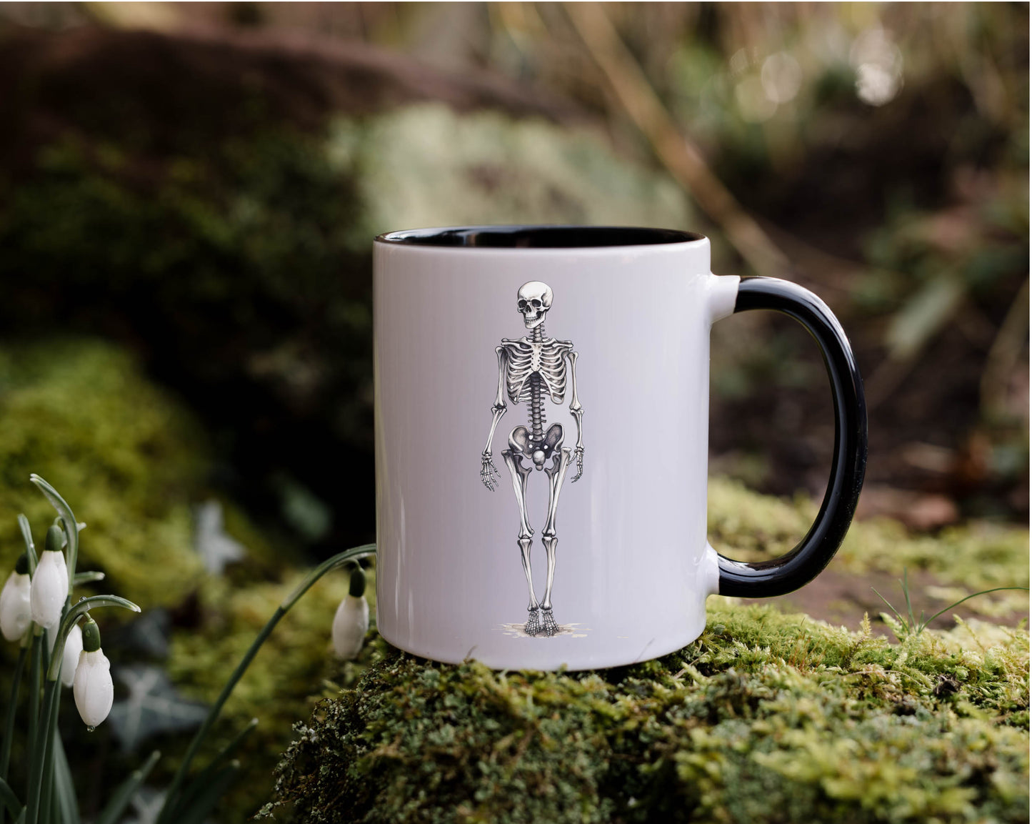 A white mug with a black interior and handle and an image of a skeleton printed on the front. The mug is set against a nature background