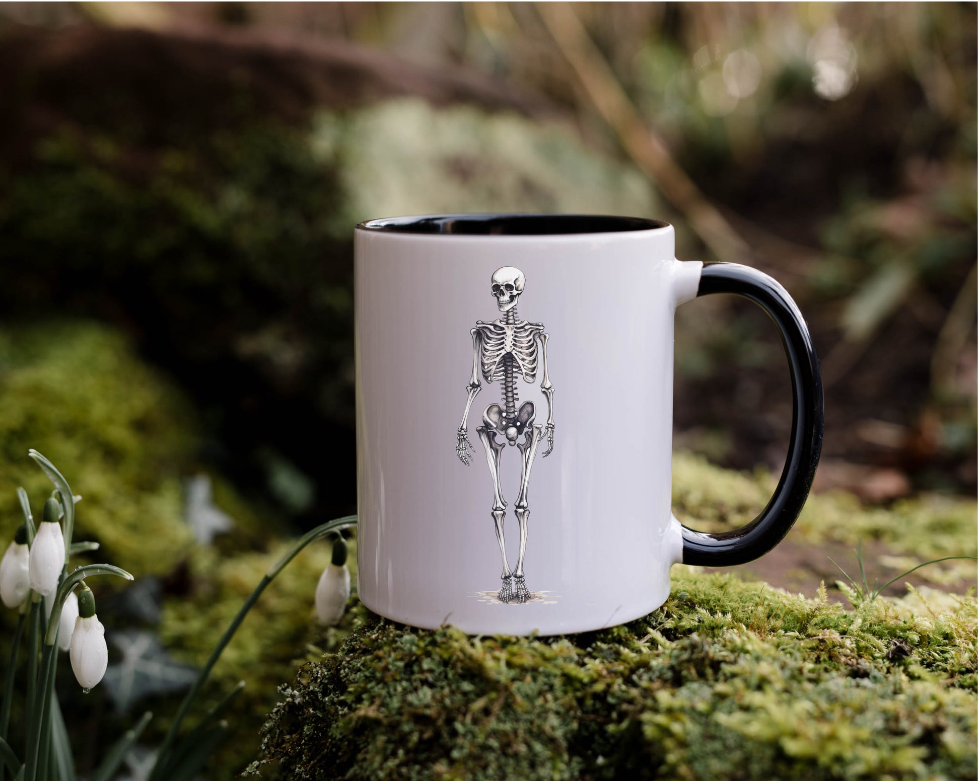 A white mug with a black interior and handle and an image of a skeleton printed on the front. The mug is set against a nature background