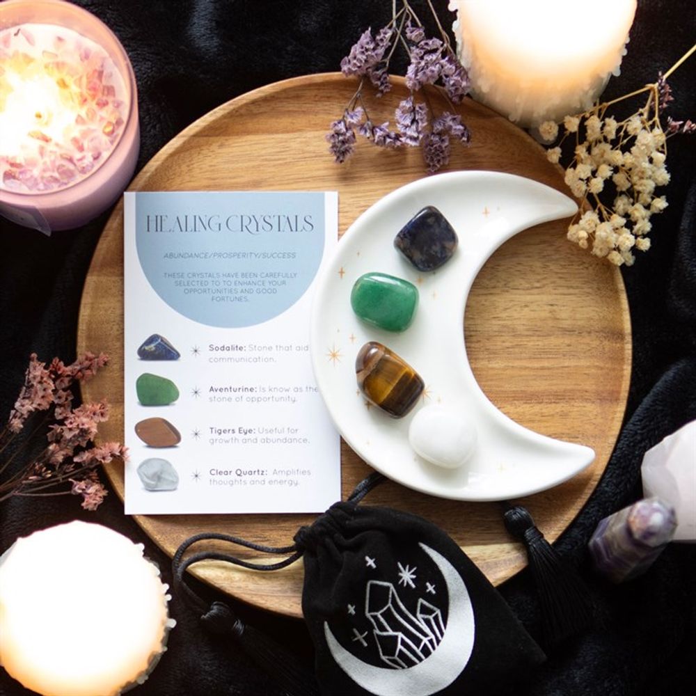 An image of a white half moon trinket dish with golden stars containing each of the 4 crystals available along with the small black velvet pouch with a moon and crystals on it. These are all placed on a wooden board with candles and herbs surrounding it