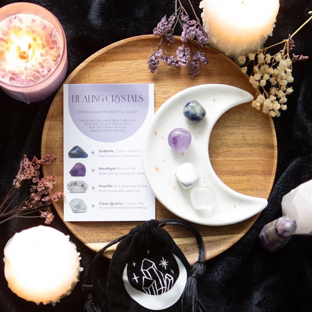 An image of a white half moon trinket dish with golden stars containing each of the 4 crystals available along with the small black velvet pouch with a moon and crystals on it. These are all placed on a wooden board with candles and herbs surrounding it