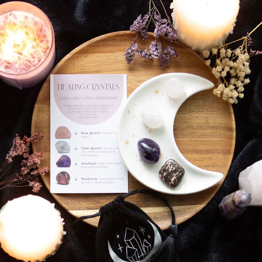 An image of a white half moon trinket dish with golden stars containing each of the 4 crystals available along with the small black velvet pouch with a moon and crystals on it. These are all placed on a wooden board with candles and herbs surrounding it